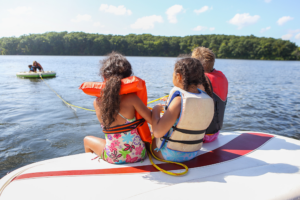 Three Children sitting on a boat wearing lifejackets, looking out onto a lake with two children on a flotation device. | Credit Soupstock on Canva
